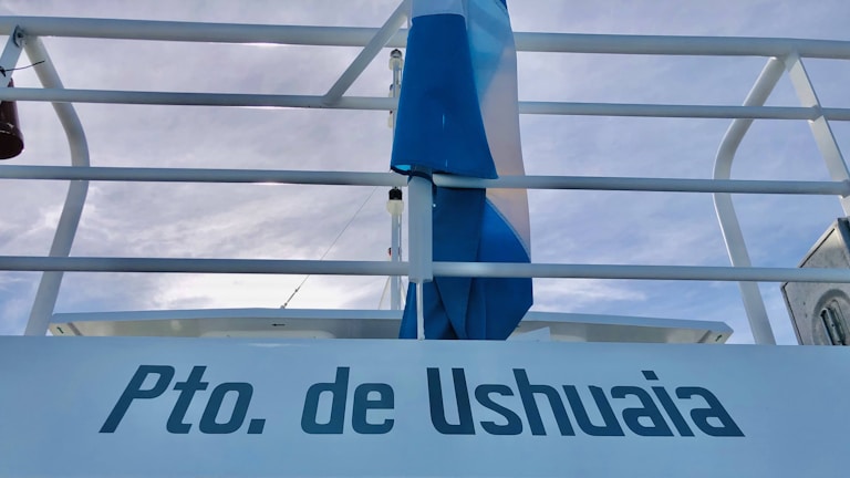 A railing with a flag in blue and white colors is visible against a cloudy sky. The text 'Pto. de Ushuaia' is prominently displayed.