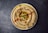 A beautifully presented plate of creamy hummus garnished with whole chickpeas, olive oil, a sprig of parsley, and a sprinkle of paprika. The hummus is arranged in a swirl pattern, with the topping centrally located, enhancing the visual appeal.
