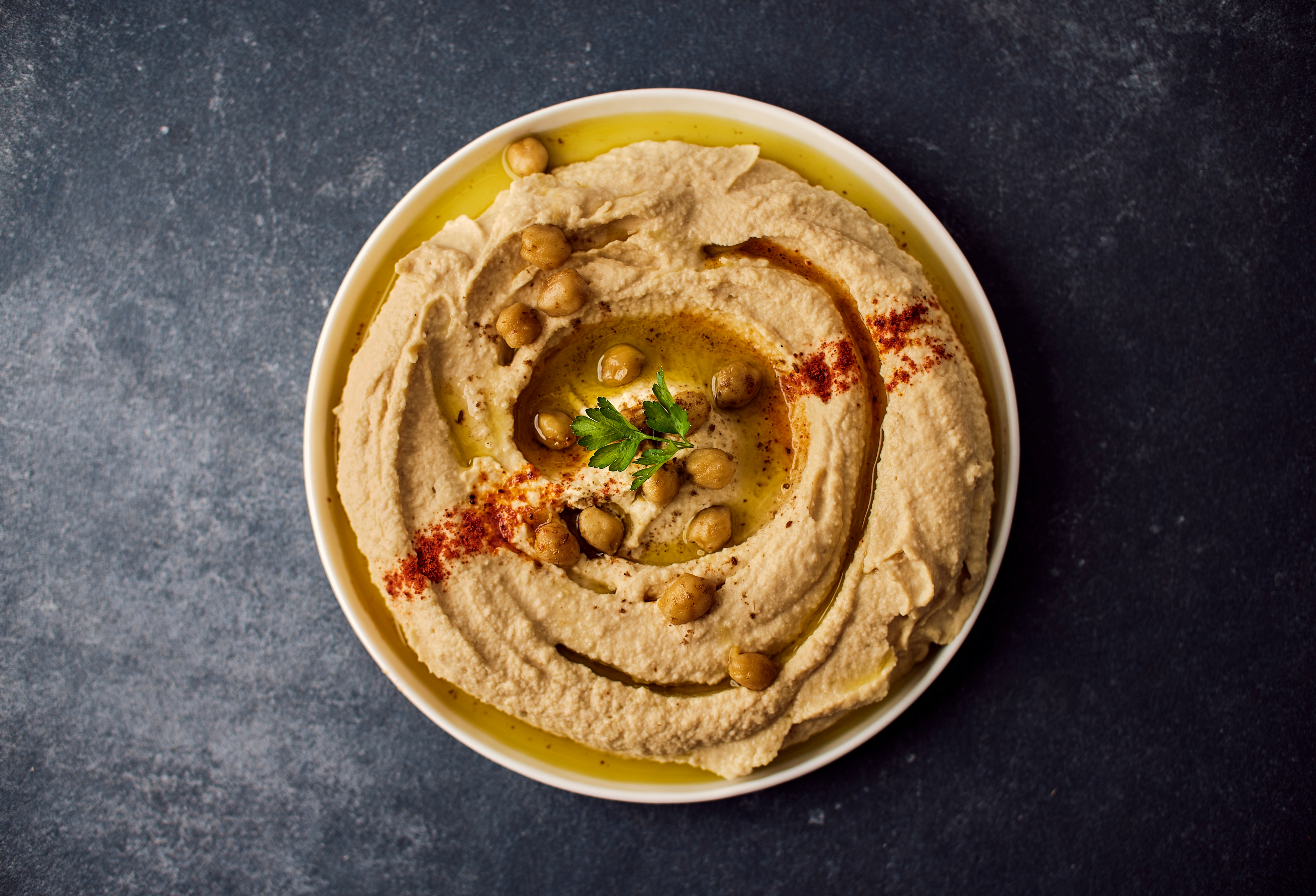 A bowl of yellow hummus drizzled with golden olive oil and topped with chick peas and red spices on a black tabletop.