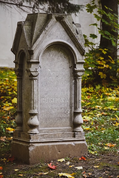 An old stone headstone stands in a grassy area, surrounded by scattered autumn leaves and partially obscured vegetation. The headstone features intricate carvings and inscriptions in a Cyrillic script, indicating its cultural or historical significance. The structure appears weathered, suggesting it has been exposed to the elements for many years.