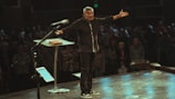 A man stands on stage with arms outstretched in front of an audience. He is dressed in dark clothing and appears to be speaking or presenting. The stage has a microphone stand, a glass lectern with an open book, and a digital tablet. The audience is visible in the background, slightly out of focus.
