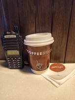 A handheld radio is placed beside a brown disposable coffee cup on a counter. The coffee cup has a white lid and features 'COFFEEHOLIC' printed in large letters. A white napkin with a 'deli2go' logo is neatly folded next to the cup. The background consists of wooden paneling.