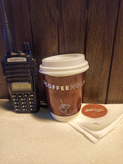 A handheld radio is placed beside a brown disposable coffee cup on a counter. The coffee cup has a white lid and features 'COFFEEHOLIC' printed in large letters. A white napkin with a 'deli2go' logo is neatly folded next to the cup. The background consists of wooden paneling.