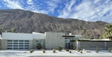 A modern, minimalist house is set against the backdrop of a rugged mountain range. The house features clean lines, a flat roof, and a front yard with desert landscaping that includes small shrubs and cacti. The sky is clear with a few scattered clouds.