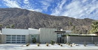 A modern, minimalist house is set against the backdrop of a rugged mountain range. The house features clean lines, a flat roof, and a front yard with desert landscaping that includes small shrubs and cacti. The sky is clear with a few scattered clouds.