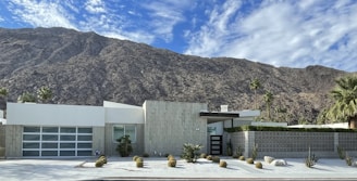 A modern, minimalist house is set against the backdrop of a rugged mountain range. The house features clean lines, a flat roof, and a front yard with desert landscaping that includes small shrubs and cacti. The sky is clear with a few scattered clouds.