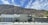 A modern, minimalist house is set against the backdrop of a rugged mountain range. The house features clean lines, a flat roof, and a front yard with desert landscaping that includes small shrubs and cacti. The sky is clear with a few scattered clouds.