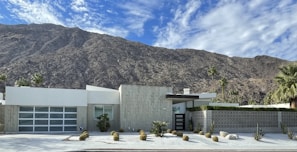 A modern, minimalist house is set against the backdrop of a rugged mountain range. The house features clean lines, a flat roof, and a front yard with desert landscaping that includes small shrubs and cacti. The sky is clear with a few scattered clouds.