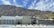 A modern, minimalist house is set against the backdrop of a rugged mountain range. The house features clean lines, a flat roof, and a front yard with desert landscaping that includes small shrubs and cacti. The sky is clear with a few scattered clouds.