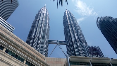 Two tall, modern skyscrapers with a distinctive architectural design and a connecting bridge between them. The buildings rise against a clear blue sky and are part of an urban setting with other high-rise structures. The sign 'Suria KLCC' is visible, indicating a commercial or shopping area.