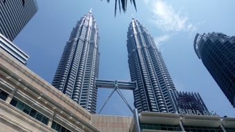 Two tall, modern skyscrapers with a distinctive architectural design and a connecting bridge between them. The buildings rise against a clear blue sky and are part of an urban setting with other high-rise structures. The sign 'Suria KLCC' is visible, indicating a commercial or shopping area.