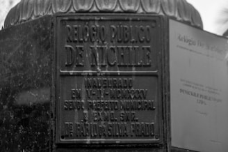 A black and white image of a public clock plaque with inscriptions in Portuguese. The text includes 'Relógio Público de Nichile' and mentions a specific inauguration date, as well as names and titles.