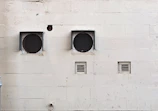 A weathered beige wall with two large circular ventilation openings covered by metal grilles. Below, there are two smaller rectangular vents. The wall has visible stains and marks, giving it an aged appearance.