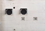 A weathered beige wall with two large circular ventilation openings covered by metal grilles. Below, there are two smaller rectangular vents. The wall has visible stains and marks, giving it an aged appearance.