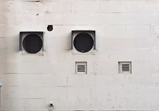 A weathered beige wall with two large circular ventilation openings covered by metal grilles. Below, there are two smaller rectangular vents. The wall has visible stains and marks, giving it an aged appearance.
