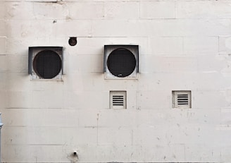 A weathered beige wall with two large circular ventilation openings covered by metal grilles. Below, there are two smaller rectangular vents. The wall has visible stains and marks, giving it an aged appearance.