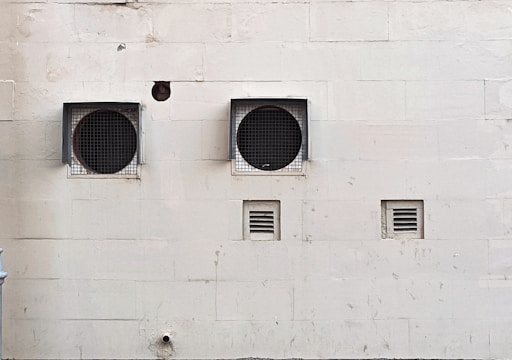 A weathered beige wall with two large circular ventilation openings covered by metal grilles. Below, there are two smaller rectangular vents. The wall has visible stains and marks, giving it an aged appearance.