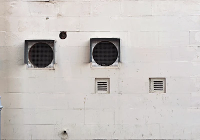 A weathered beige wall with two large circular ventilation openings covered by metal grilles. Below, there are two smaller rectangular vents. The wall has visible stains and marks, giving it an aged appearance.