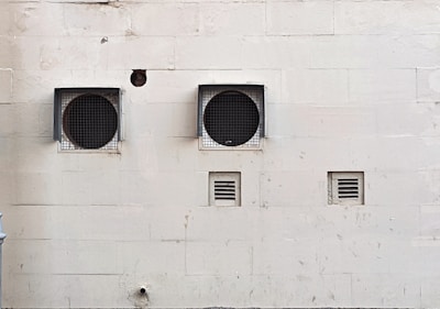 A weathered beige wall with two large circular ventilation openings covered by metal grilles. Below, there are two smaller rectangular vents. The wall has visible stains and marks, giving it an aged appearance.