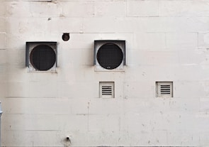 A weathered beige wall with two large circular ventilation openings covered by metal grilles. Below, there are two smaller rectangular vents. The wall has visible stains and marks, giving it an aged appearance.