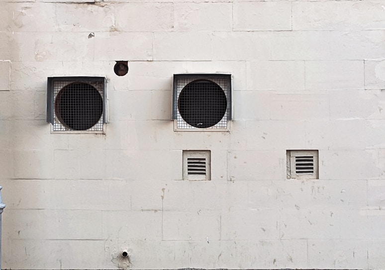 A weathered beige wall with two large circular ventilation openings covered by metal grilles. Below, there are two smaller rectangular vents. The wall has visible stains and marks, giving it an aged appearance.