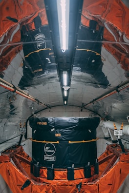 A highly reflective and metallic interior with structured walls and ceiling, housing a large black and orange module. The module has visible branding and labels indicating Intelsat and Hughes. The environment appears technical and industrial, possibly inside a spacecraft or satellite.