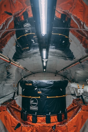 A highly reflective and metallic interior with structured walls and ceiling, housing a large black and orange module. The module has visible branding and labels indicating Intelsat and Hughes. The environment appears technical and industrial, possibly inside a spacecraft or satellite.