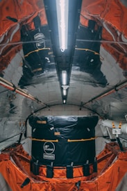 A highly reflective and metallic interior with structured walls and ceiling, housing a large black and orange module. The module has visible branding and labels indicating Intelsat and Hughes. The environment appears technical and industrial, possibly inside a spacecraft or satellite.