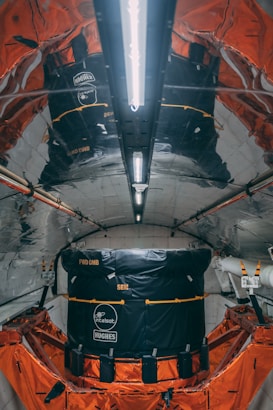 A highly reflective and metallic interior with structured walls and ceiling, housing a large black and orange module. The module has visible branding and labels indicating Intelsat and Hughes. The environment appears technical and industrial, possibly inside a spacecraft or satellite.