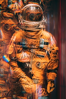 A vibrant orange space suit is displayed, complete with a helmet reflecting light and creating an array of colorful patterns. The suit bears various patches, including an American flag and a mission badge, suggesting its use in NASA missions.