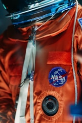 An astronaut's suit with an orange fabric adorned with a NASA patch. The suit features metallic clasps and a circular vent. The focus is on the upper body of the suit, with a helmet visible at the top.