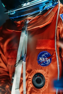An astronaut's suit with an orange fabric adorned with a NASA patch. The suit features metallic clasps and a circular vent. The focus is on the upper body of the suit, with a helmet visible at the top.