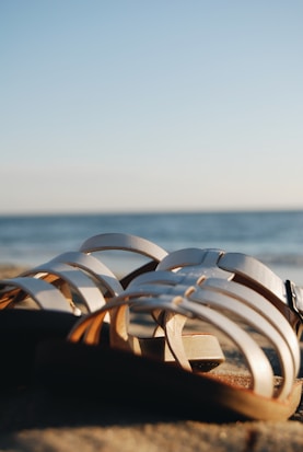 A pair of white sandals lies on a sandy beach near the water's edge. The sea is gently visible in the background, and the sky is clear with hints of blue.