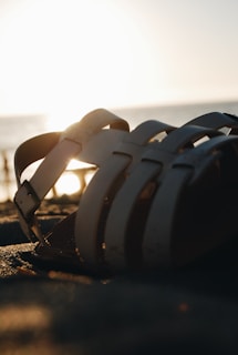 A sandal lying on the sand at the beach during sunset. The sun is low in the sky, casting a warm glow over the scene and creating long shadows from the sandal straps. The background features the blurred horizon with a calm sea.