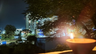 A nighttime scene features a lit diya in the foreground, with its warm glow contrasting against the dark. Tall buildings with lit windows and neon lights are visible in the background, partially obscured by leafy tree branches.