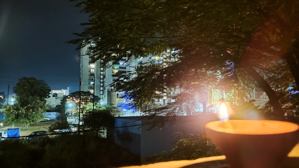 A nighttime scene features a lit diya in the foreground, with its warm glow contrasting against the dark. Tall buildings with lit windows and neon lights are visible in the background, partially obscured by leafy tree branches.