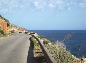 A coastal road curves alongside the blue sea, with a steep rocky cliff on the left and greenery lining the right edge. A small red and white chapel is visible in the distance, perched near the cliff edge.