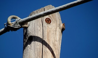 A wooden utility pole with visible knots and cracks stands against a clear blue sky. A metal cable is wrapped around the top, securely attached to the pole.