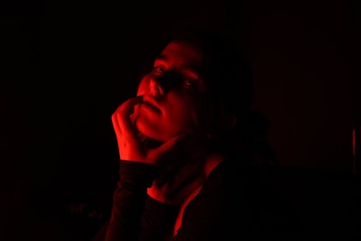 A person is partially illuminated by red light, creating a dramatic and shadowy effect. The light highlights their face and hand against a dark background, with soft red tones casting dynamic shadows.