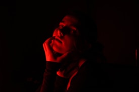 A person is partially illuminated by red light, creating a dramatic and shadowy effect. The light highlights their face and hand against a dark background, with soft red tones casting dynamic shadows.