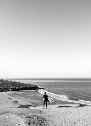 A person in a wetsuit stands on a sandy cliff overlooking the ocean while holding a surfboard. The shoreline curves around rocky terrain, and gentle waves lap against the beach. The sky is clear, creating a serene and spacious feeling.