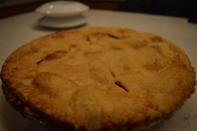 A freshly baked pie with a golden-brown crust sits on a white surface. In the background, there is a blurred view of a white dish and lid.