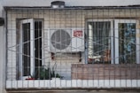 A balcony enclosed with metal bars featuring a white air conditioning unit mounted on a brick wall. Two glass doors with white frames, one open and one closed, are visible. Several potted plants and a red bowl are placed on the windowsill inside the balcony.