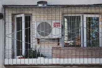 A balcony enclosed with metal bars featuring a white air conditioning unit mounted on a brick wall. Two glass doors with white frames, one open and one closed, are visible. Several potted plants and a red bowl are placed on the windowsill inside the balcony.