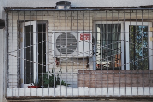A balcony enclosed with metal bars featuring a white air conditioning unit mounted on a brick wall. Two glass doors with white frames, one open and one closed, are visible. Several potted plants and a red bowl are placed on the windowsill inside the balcony.