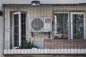 A balcony enclosed with metal bars featuring a white air conditioning unit mounted on a brick wall. Two glass doors with white frames, one open and one closed, are visible. Several potted plants and a red bowl are placed on the windowsill inside the balcony.