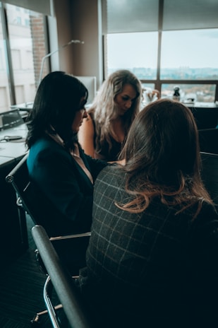 A diverse group of professional women engaged in a thoughtful leadership discussion around a conference table with subtle purple and teal accents in the background.