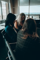 Three women are seated around a table in a modern office setting, engaged in a discussion or meeting. The room has large windows with a view of buildings outside and chairs that have a sleek, professional design. The lighting is subdued, creating a calm atmosphere.