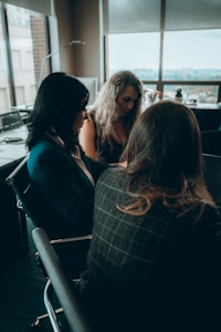 Three women are seated around a table in a modern office setting, engaged in a discussion or meeting. The room has large windows with a view of buildings outside and chairs that have a sleek, professional design. The lighting is subdued, creating a calm atmosphere.
