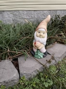 A small garden gnome with a white beard and red hat is sitting among green grass and plants. It holds a green object resembling a tree. The gnome is placed on concrete blocks with a background of a stone wall and siding.